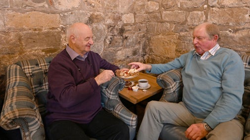 Two men sat in arm chairs with a pot of tea and a scone with cream and jam